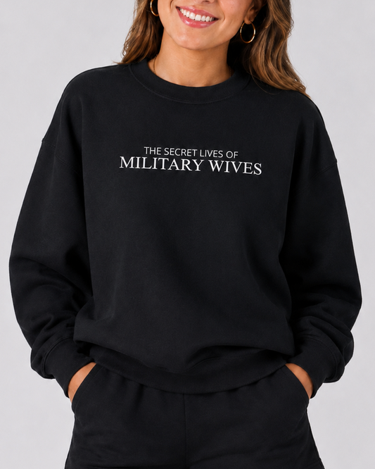 A woman wearing a black crewneck sweatshirt with the text 'THE SECRET LIVES OF MILITARY WIVES' in white, paired with black shorts, against a solid gray studio background.